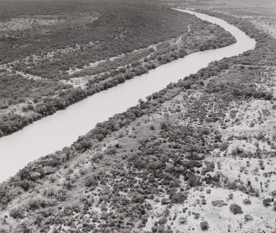 A black-and-white aerial photograph of a river running through a scrubby landscape.
