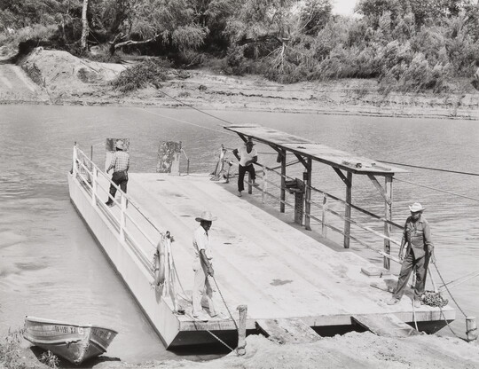 A black-and-white photograph of several people standing on a docked wooden ferry; some hold the ropes that tether it to shore.