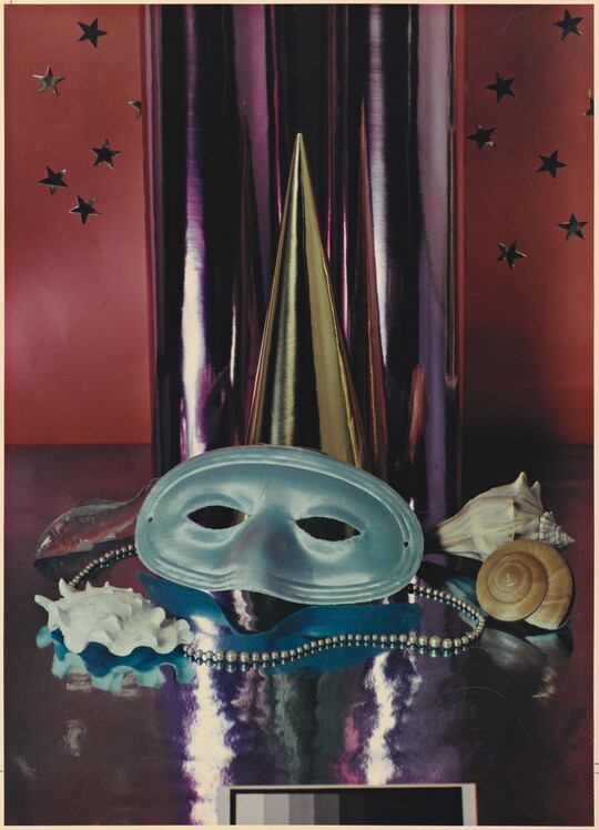 A color photograph of a simple party mask, Mardi Gras beads, and seashells in front of a reflective silver cylinder.