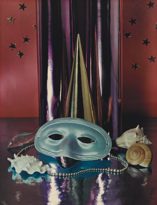 A color photograph of a simple party mask, Mardi Gras beads, and seashells in front of a reflective silver cylinder.