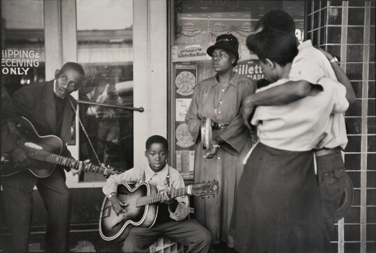 A black-and-white photograph of Black adults and boy playing instruments in front of a storefront while a Black couple watches.