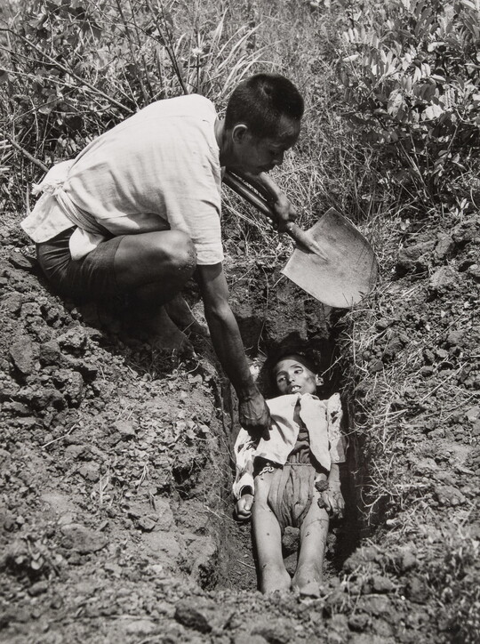 A black-and-white photograph of an Asian man holding a shovel crouched down next to a freshly dug grave containing a emaciated body.