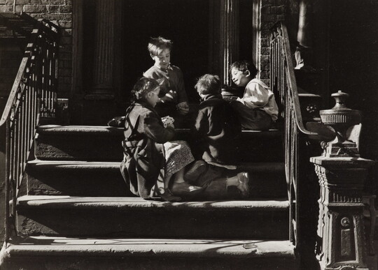 A black-and-white photograph of four children playing together on concrete steps with iron railings that cast striped shadows across them.