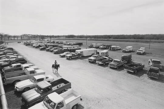 A black-and-white photograph of a single rider on horseback amid rows of parked cars and trucks.