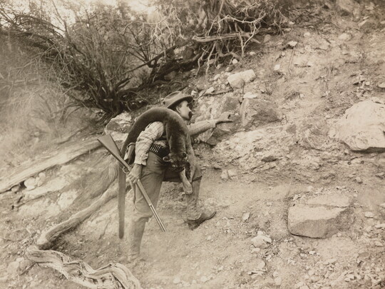 A black-and-white photograph of a White man climbing a rocky ridge holding a rifle with a mountain lion carcass over his shoulder.