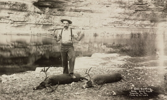 A black-and-white photograph of a White man racking a rifle across his shoulders while standing over two buck carcasses next to a creek.