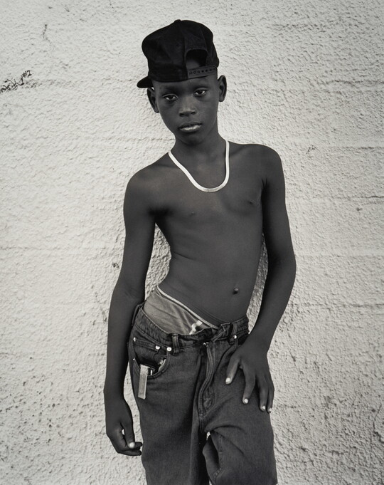 A black-and-white portrait photograph of a Black teenage boy wearing a backward baseball cap, a necklace, no shirt, and loose fitting jeans against a concrete wall.