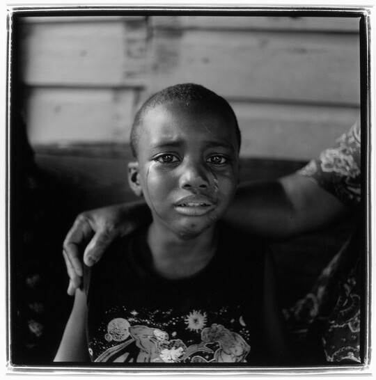 A black-and-white photograph of a young Black child with tears streaming down their face and someone's arm around their shoulders.