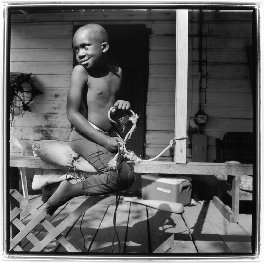 A black-and-white photograph of a young Black boy straddling the railing of a porch pretending to ride a horse.