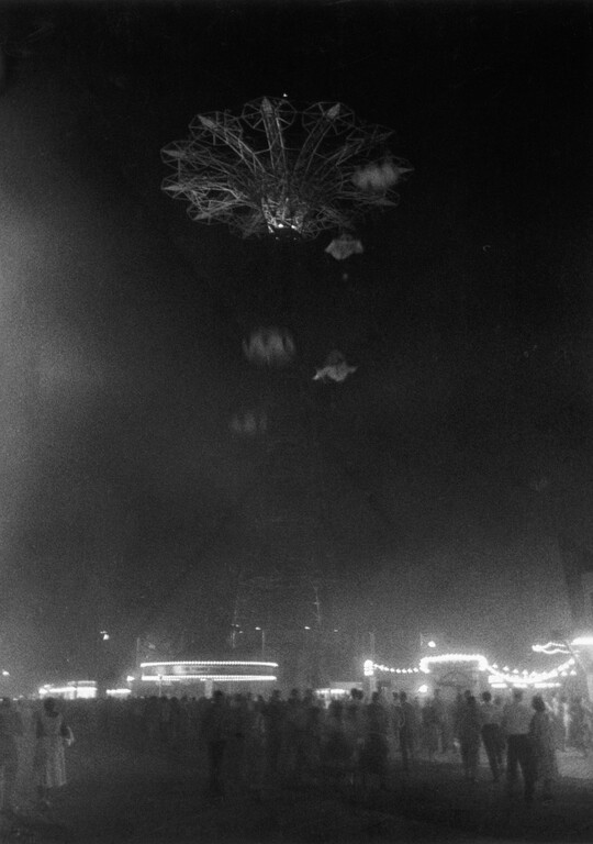 A black-and-white photograph looking up at a tall ride on festival grounds at night.
