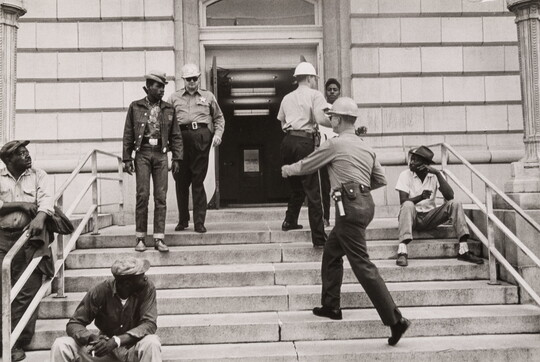 A black-and-white photograph of Black men on steps, two holding protest signs, and two White police officers approaching them, one with a baton.