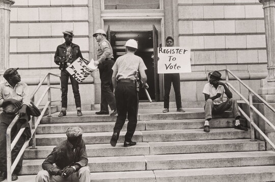 A black-and-white photograph of Black men on steps, two holding protest signs, and two White police officers approaching them, one with a baton.