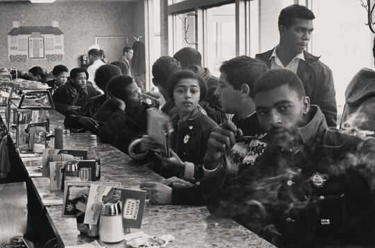 A black-and-white photograph of mostly young Black people and one White person at a crowded diner counter, talking and smoking.