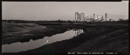 A black-and-white panoramic photograph of a wide, flat landscape with a river reflecting part of a city skyline in the distance.