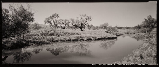 A panoramic black-and-white photograph of a calm body of water winding through a scrubby landscape.