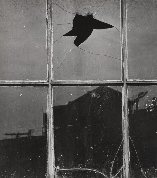 A black-and-white photograph of dirty and cracked windowpanes, one with a hole through it, and a silhouette of a building reflected in the glass.
