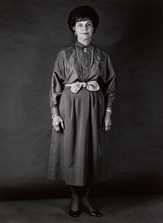 A black-and-white portrait photograph, in full length, of a middle aged White woman wearing a long-sleeved dress with a light-colored bow at her waist.