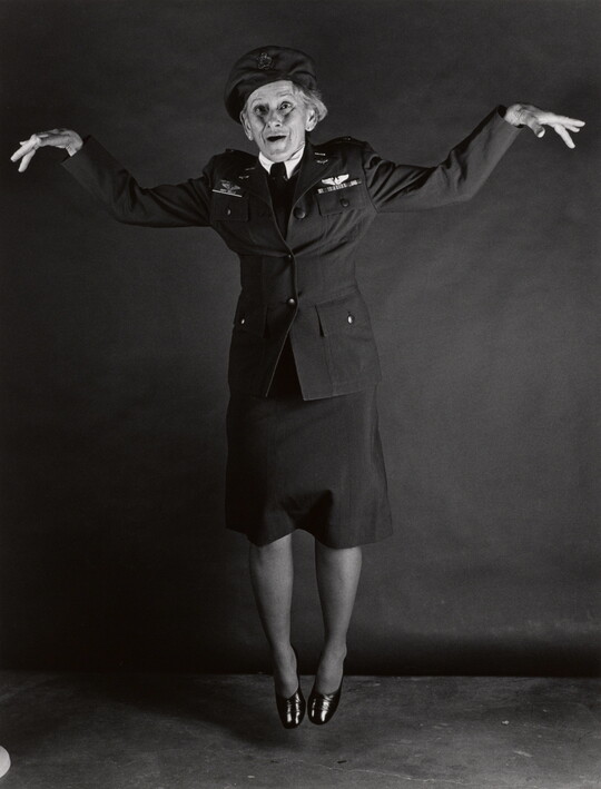 A black-and-white portrait photograph, in full length, of an older White woman wearing military uniform, jumping and striking a flying-pose with her arms.
