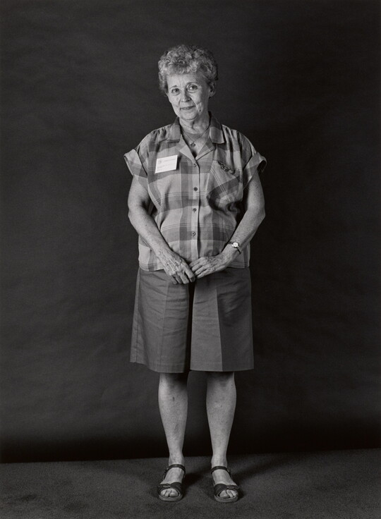 A black-and-white portrait photograph, in full length, of an older White woman with a shy smile wearing a short-sleeved plaid shirt and culotte shorts.