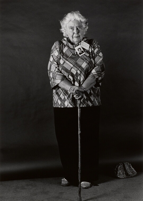 A black-and-white portrait photograph, in full length, of a white-haired, elderly White woman wearing a plaid shirt, long dark pants, and resting her hands on a cane.