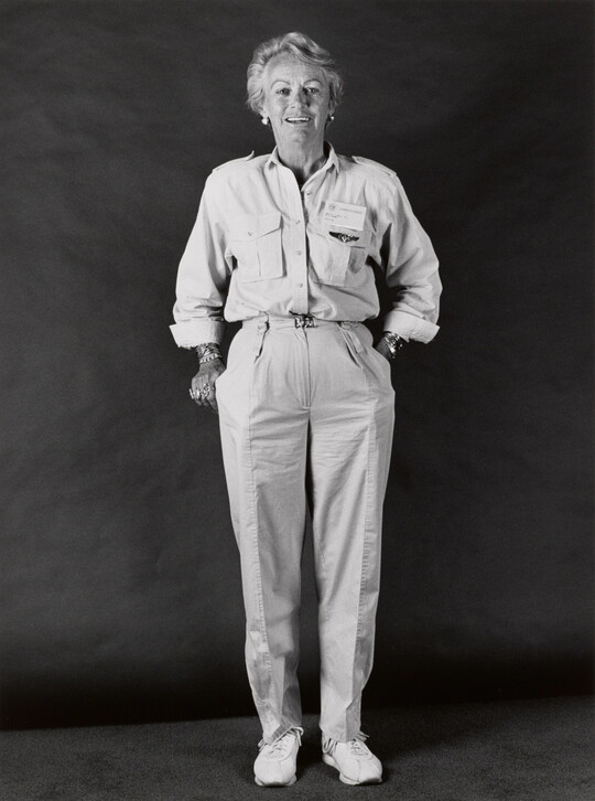 A black-and-white portrait photograph of a smiling middle-aged White woman with short hair wearing a button-down shirt with a name badge above the breast pocket, light colored pants, and sneakers.