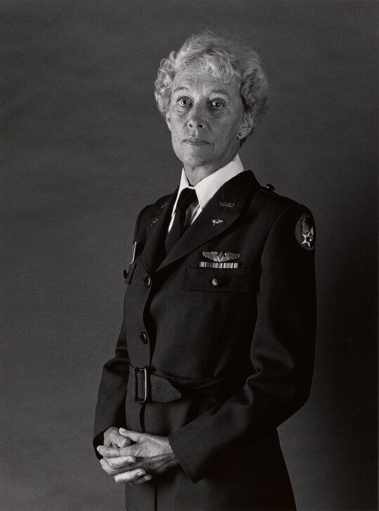 A black-and-white portrait photograph of a middle-aged White woman with curly hair wearing a WASP military uniform and her hands folded in front of her.