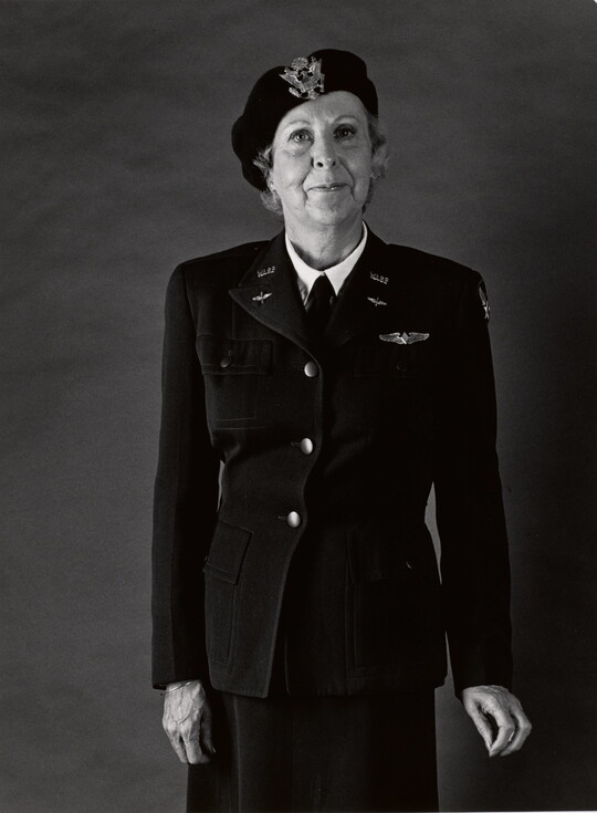 A black-and-white portrait photograph of an older White woman wearing a beret with an insignia on the front, a WASP military uniform, and holding her hands at her sides.