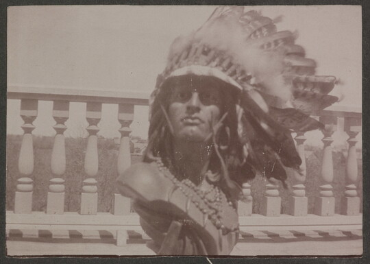 A sepia-toned photograph of a male Native American sculpture bust wearing a real feathered headdress and beads around the neck.