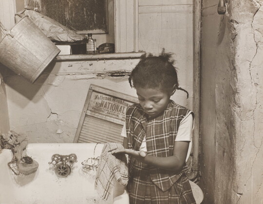 A black-and-white photograph of a young Black girl wearing a plaid vest and skirt washing her hands with a cloth in front of a bathtub.