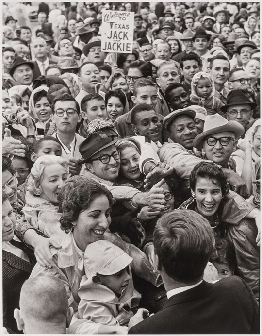 A black-and-white photograph of President Kennedy from the back facing a large crowd that is reaching up to touch his hand.