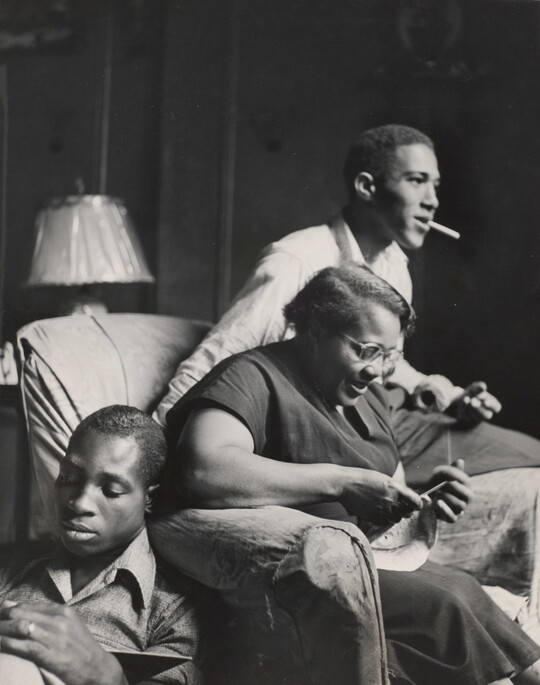 A black-and-white photograph of a Black woman knitting in an armchair, a young Black man smoking on the arm of her chair, and a young Black man seated on the floor reading.