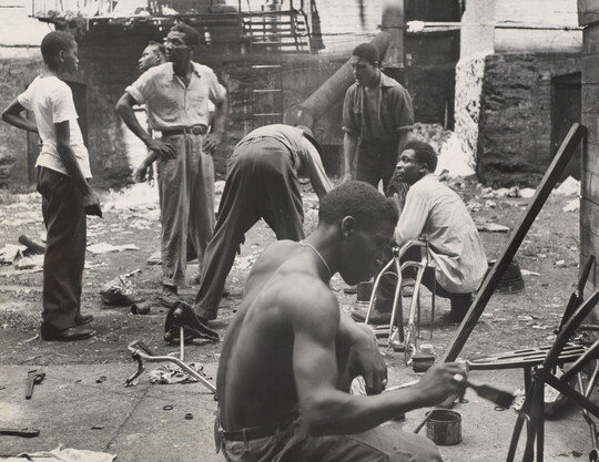 A black-and-white photograph of group of Black men talking and working in a rundown space littered with debris.