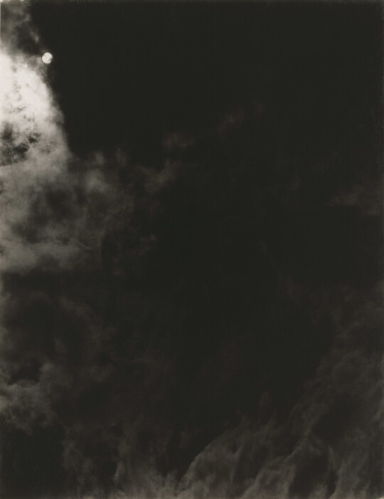 A black-and-white photograph of a dark, cloudy sky with the sun or moon shining through clouds.