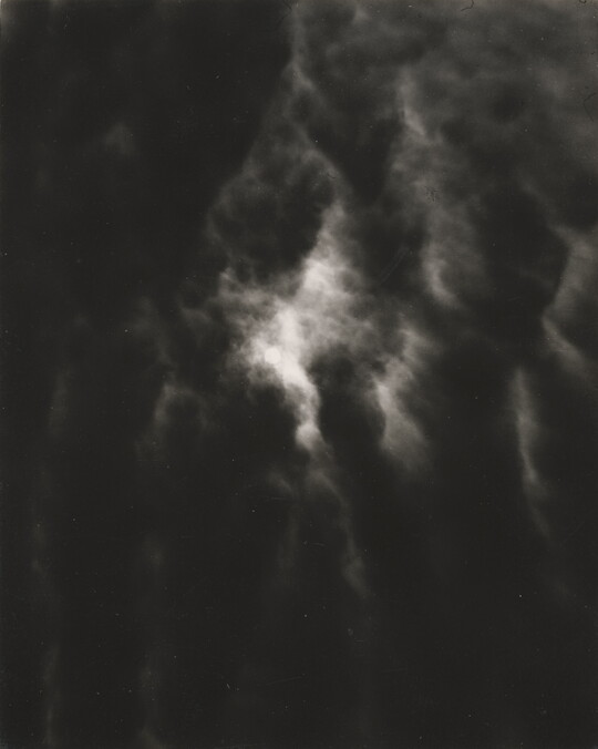 A black-and-white photograph of a dark, cloudy sky with light shining through the clouds.