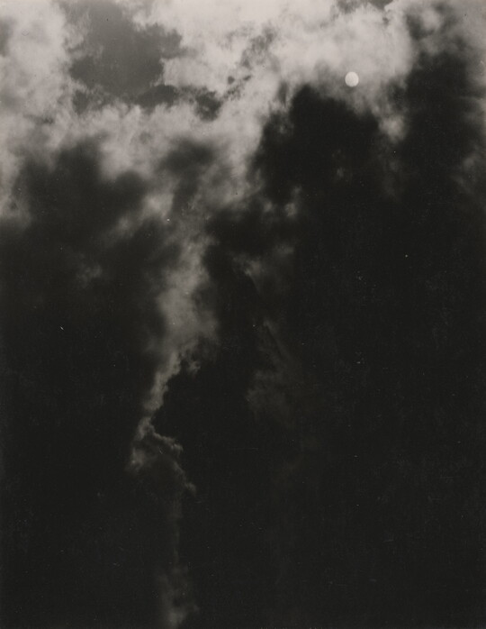 A black-and-white photograph of a dark, cloudy sky with the sun or moon shining through clouds.