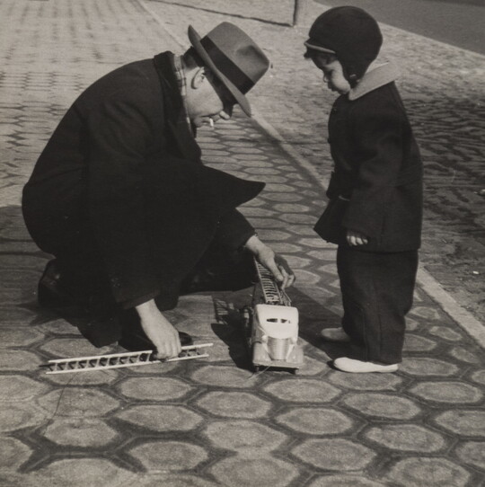 A black-and-white photograph of a small boy bundled in winter clothing watching as a man in a fedora crouches down to reach toward a toy fire truck at the boy's feet.