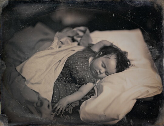 A black-and-white photograph of a White baby sleeping on their back, dark hair sticking to their head, in a bed with one hand above their head and the other at their side.