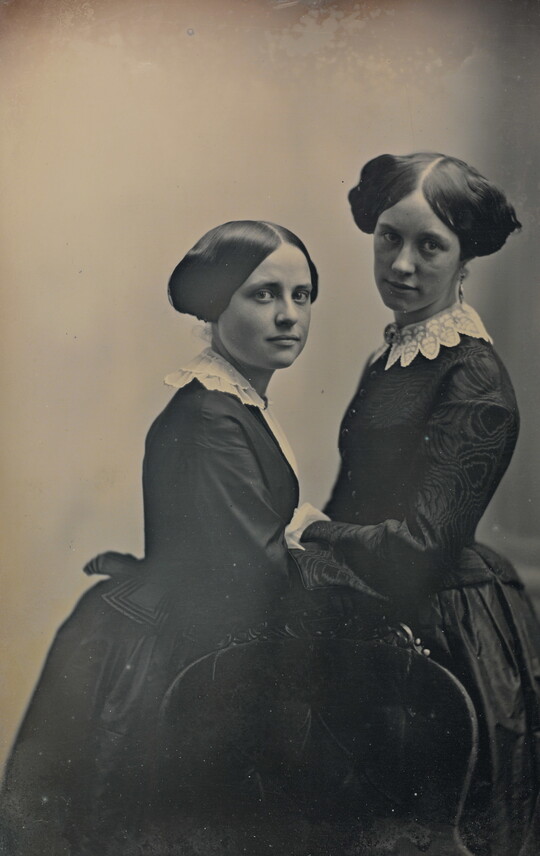 A black-and-white portrait photograph of two young White girls facing each another in period clothing and hairstyles.