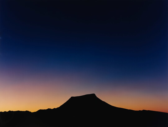 A color photograph of a sky that goes from almost black to blue to gold where it meets the silhouette of a flat topped mountain.