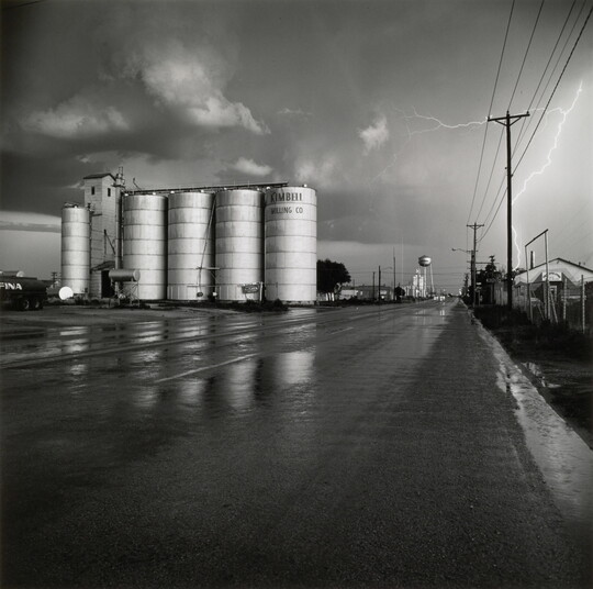 A black-and-white photograph of four grain elevators to the left of a wet road, a water tower in the far distance, and a flash of lighting behind power lines on the right.