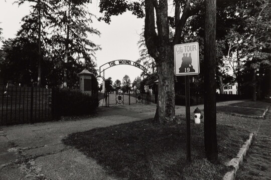 A black-and-white photograph taken from the street of a wrought iron gate a the entrance to a cemetery.