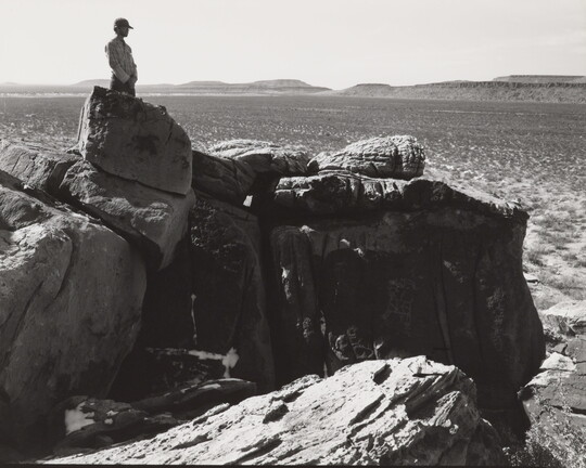 A black-and-white photograph of a man standing on a rocky cliff overlooking a wide canyon.