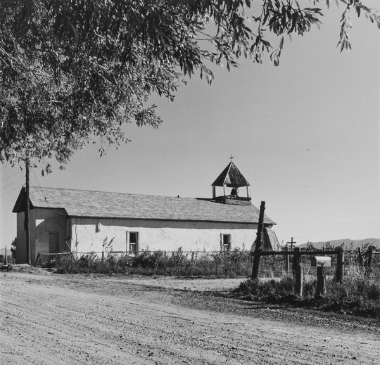 A black-and-white photograph of a long, narrow white building with a small steeple on a dusty road behind a fence.