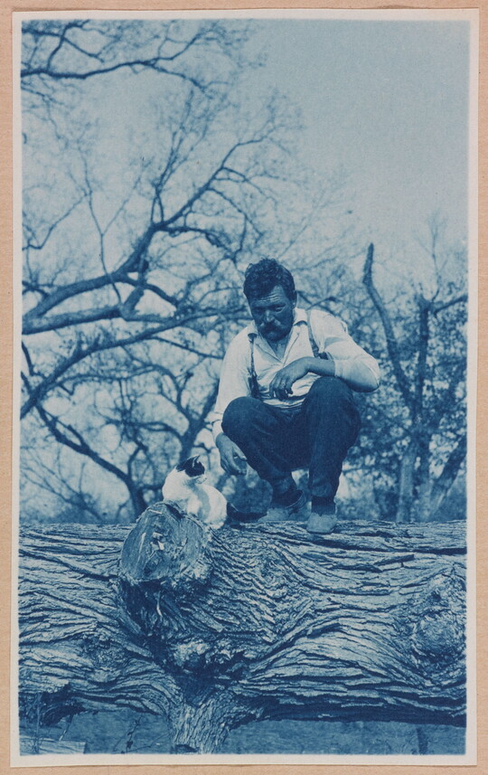 A black-and-white photograph of a White man squatting on a large log next to a white-and-black cat.