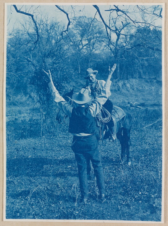 A black-and-white photograph of a man in a cowboy hat standing with both hands raised up in front of a smiling man wearing a cowboy hat on horseback.