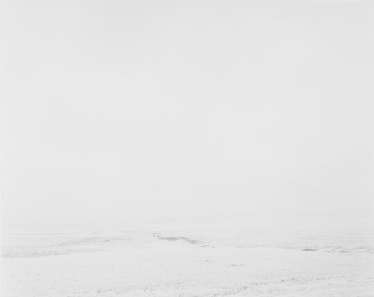 A black-and-white photograph of a white, snow-covered landscape with scattered hints of rocks or vegetation against a white sky.