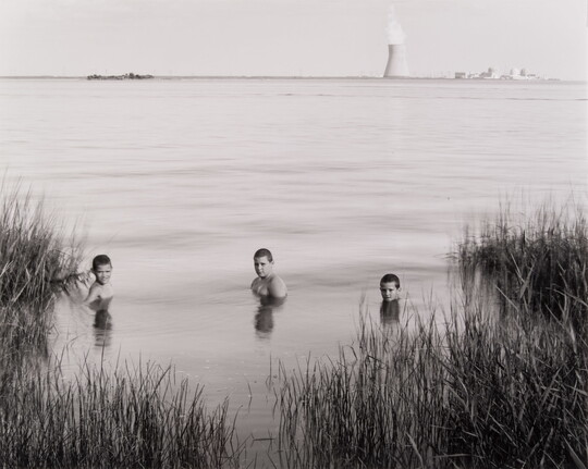 A black-and-white photograph of three White boys submerged to their shoulders in a lake; on the opposite shore in the background, a nuclear reactor puffs steam into the sky.