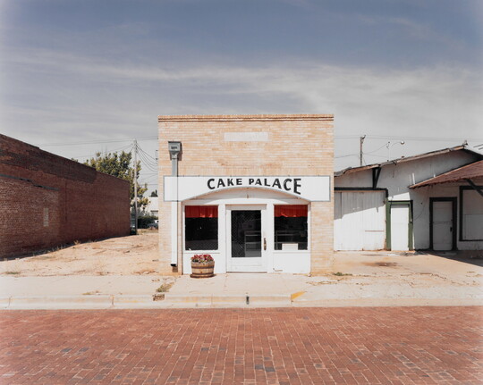 A color photograph of a tan brick building with a sign that reads "Cake Palace" above the door on a red brick road.