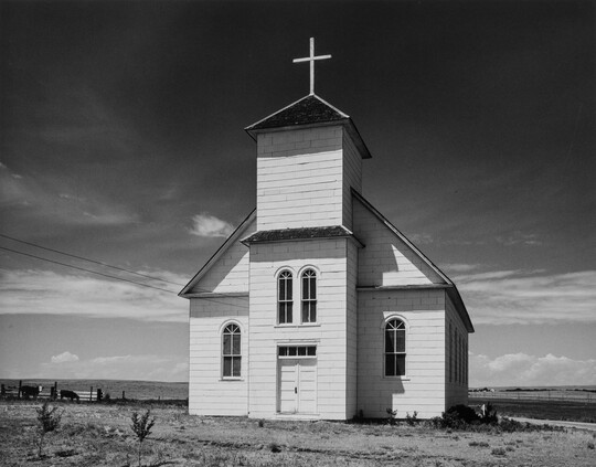 A black-and-white photograph of a small, white clapboard church, viewed from the front, under dark clouds.