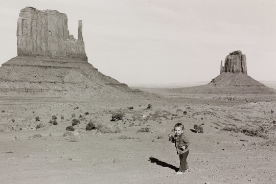 A black-and-white photograph of a toddler in front of tall buttes rising from the desert ground.